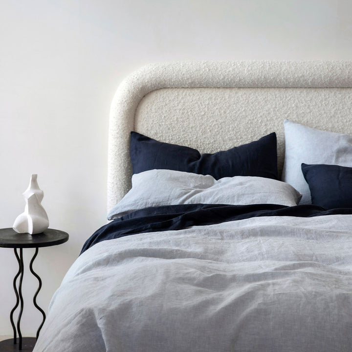 A cream bed dressed in Navy and Sky bed linen, styled with a modern black bedside table and small white ornament