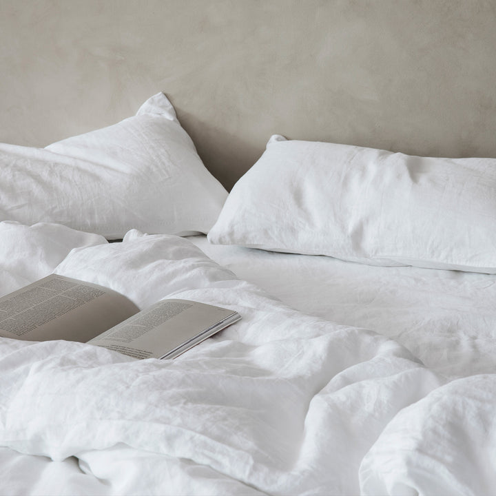 A bed in front of a beige textured wall. The bed is dressed in white linen, and has an open book resting on top.
