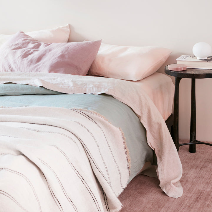A bed dressed in a Sheet Set in Blush with a Pillowcase in Dusk, a Duvet Cover in Sage and a Mira Ana Throw. A black side table holds a small lamp and a book. Sizes: Queen, Kiing