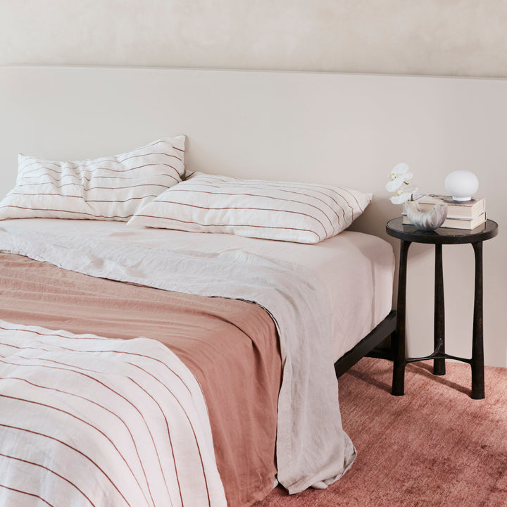 A bed dressed in Cedar Stripe, Fawn, Smoke Grey and Blush bed linen, styled with a black bedside table, small vase and a couple of books. Sizes: Queen, King