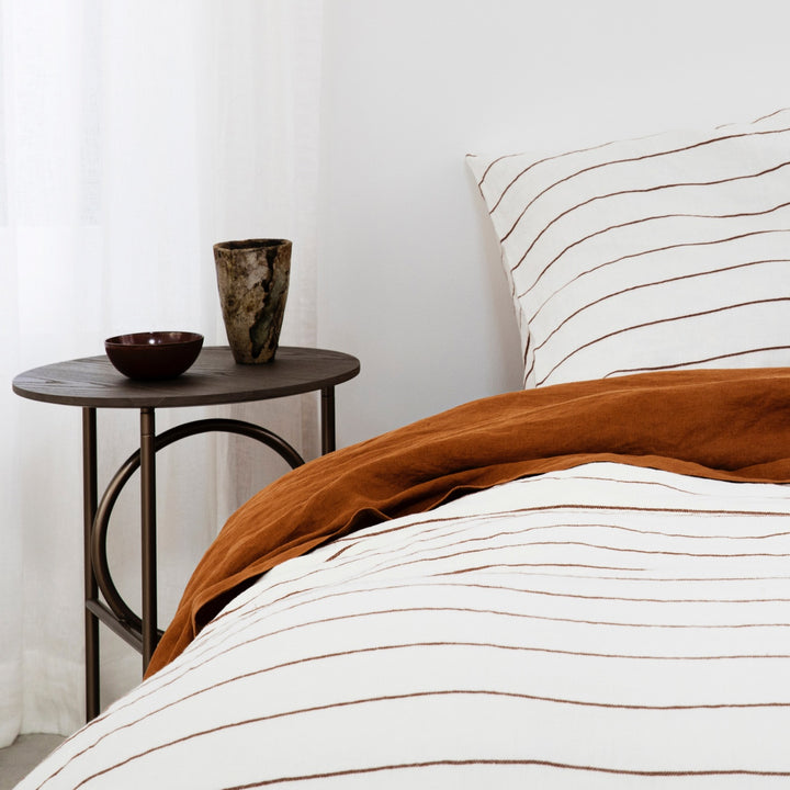 A close up of a bed dressed in Cedar Stripe and Cedar bed linen, styled with a modern bedside table, vase and bowl. Sizes: Queen, King