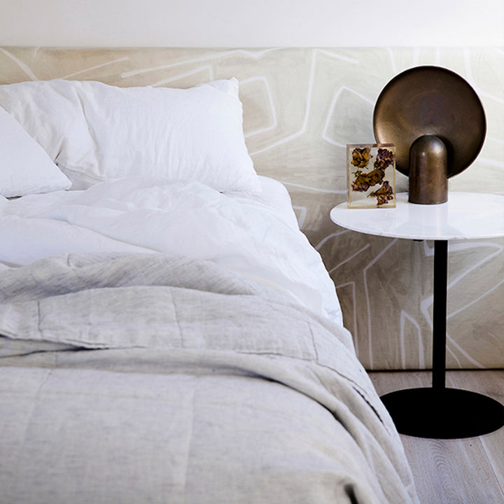 A bed dressed in White bedlinen, with a Pinstripe Quilt Cover, is styled against a printed bedhead and a side table with brass objects.