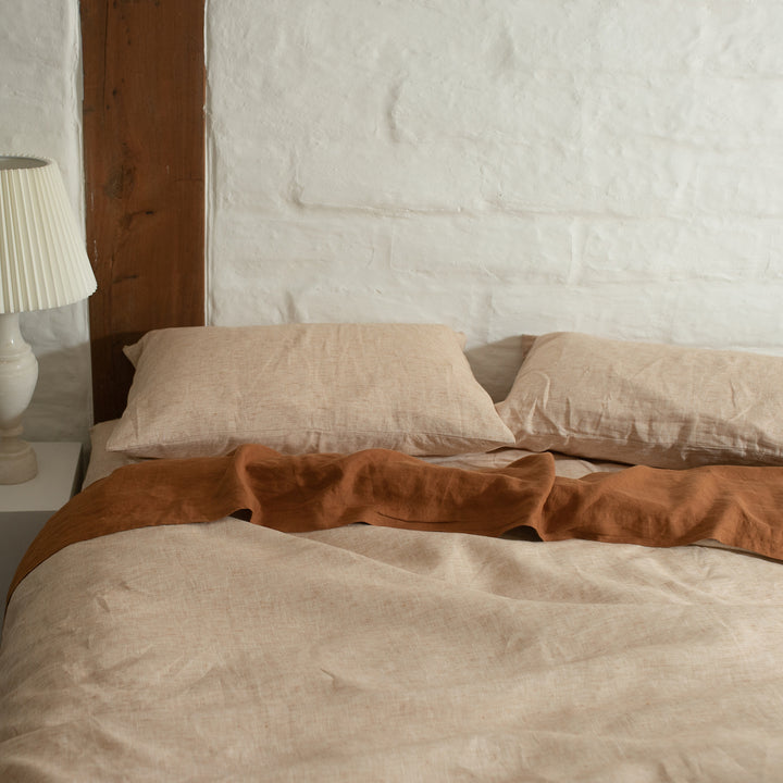A bed dressed in Cinnamon bedlinen with a Flat sheet in Cedar, styled with a side table and white lamp.