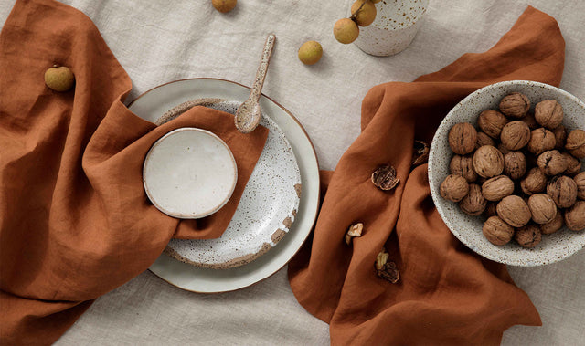 A table setting styled with a Linen Tablecloth in Natural. On top of the tablecloth are Linen Table Napkins in Cedar styled with speckled white ceramic crockery and a bowl of walnuts.