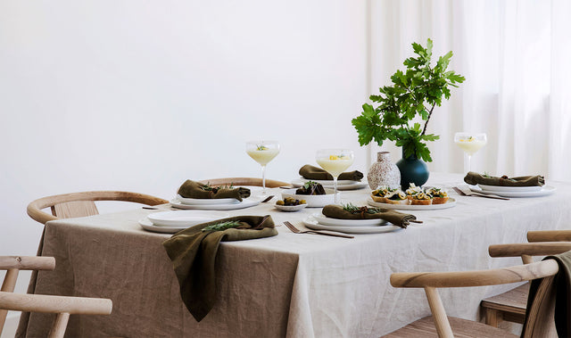 A table setting with white crockery and dark cutlery styled with a Linen Tablecloth in Natural and Linen Table Napkins in Olive amongst a plate of entrees, cocktails and 2 vases, one of which contains some branches.