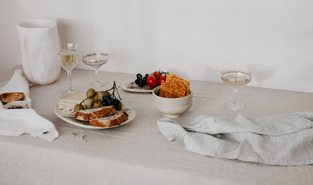 A table setting styled with a Linen Table Cloth in Natural and Linen Table Napkins in White and Pinstripe. On top of the table cloth are some cocktail and wine glasses and plates and bowls containing various fruits, bread, crackers and cheese. 