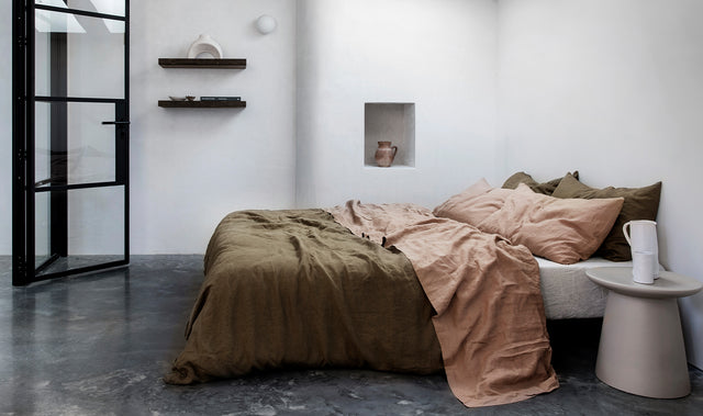 A bed in a room with white walls and dark textured floors. The bed is styled with a Linen Duvet Cover Set in Olive, Linen Flat Sheet with Border and Set of 2 Linen Pillowcases in Fawn and a Linen Fitted Sheet in Natural. Next to the bed is a gray side table with a ceramic white jug and cup on it. 