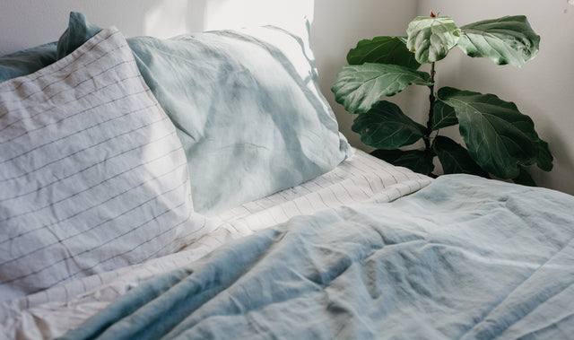 A bed styled with a Linen Duvet Cover Set in Sage and a Linen Sheet Set with Pillowcases in Pencil Stripe. Next to the bed is a fiddle leaf fig plant.