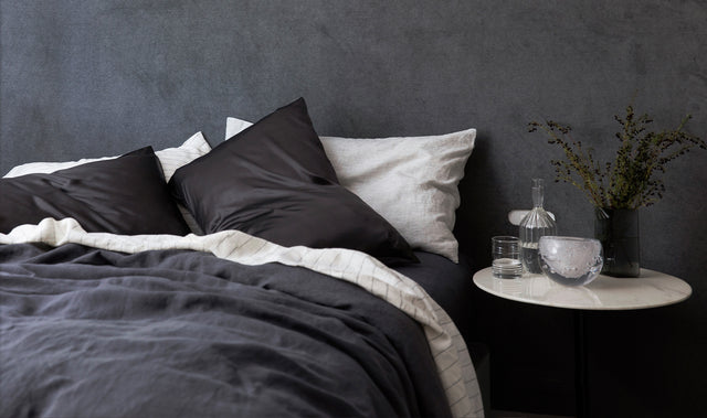 A bed in front of a dark gray textured wall. The bed is styled with a Slate Duvet Cover and Fitted Sheet, Pencil Stripe Flat Sheet, Slate Silk Linen Flip Pillowcases and Pinstripe Pillowcases. Next to the bed is a white marble bedside table that has glassware pieces and some dark florals in a vase.