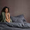 A model is sitting on bed that is styled with CULTIVER Slate linen. This includes a Duvet Cover, Fitted Sheet and Pillowcases. The model has dark skin and dark curly hair, and is draped in a Forest Waffle Linen Throw. Next to the bed is a gray bedside table with a lamp. 