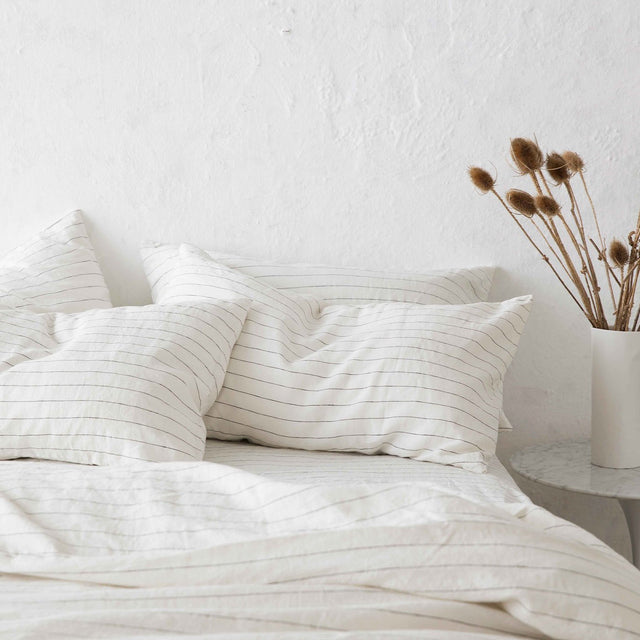 Set of 2 Linen Pillowcases and Linen Duvet Cover in Pencil Stripe on a bed. Next to the bed is a marble bedside table with a white vase containing brown florals.