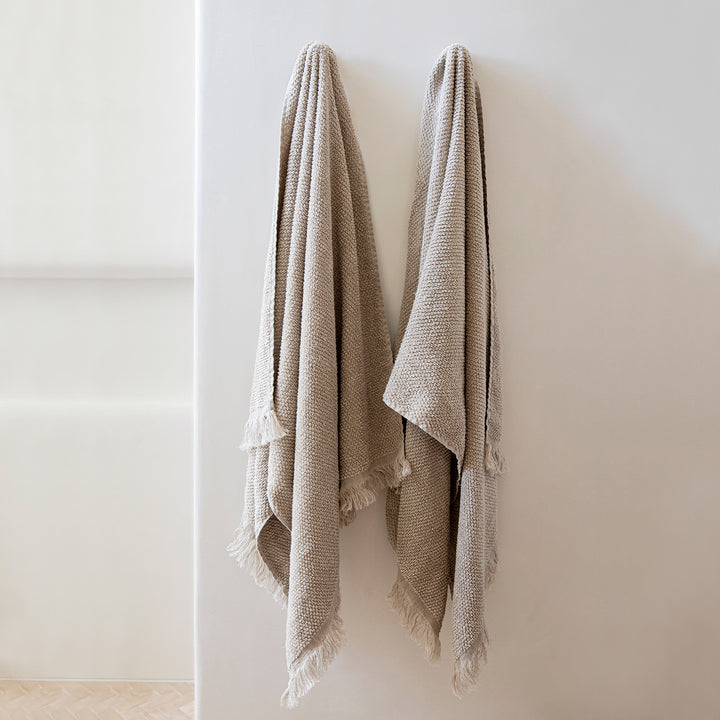Two Pure Linen Bath Towels in Natural hanging on hooks on a white wall.