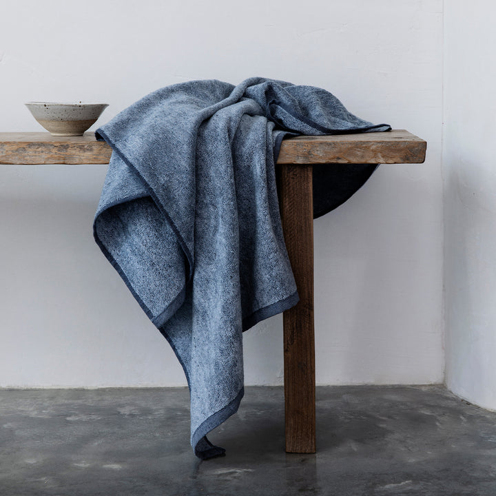 A denim bath towel draped on a bench. Sizes: Bath Towel Bundle - 28" x 55", Bath Sheet Bundle - 35" x 69"