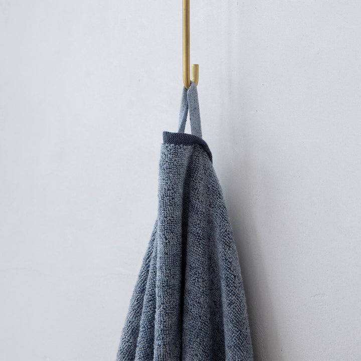 A close up photo of a Denim bath towel hanging on hook. Sizes: Bath Towel - 28" x 55", Bath Sheet - 35" x 69"