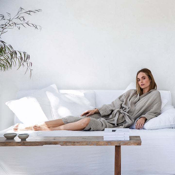 A woman lounging on a white sofa is wearing a Linen Waffle Robe in Natural. Sizes: S/M and M/L